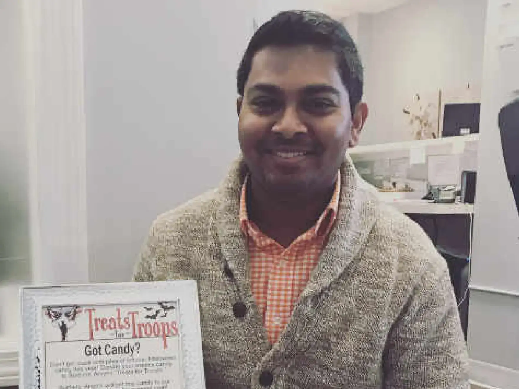 Man holding "Treats for Troops" poster in dental office setting, smiling at camera.