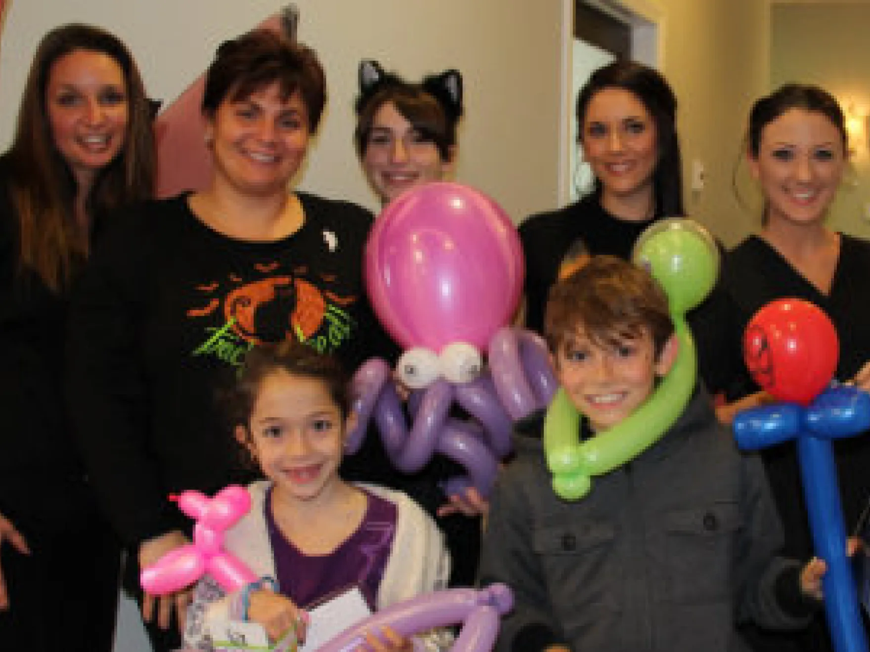 Group of women and children with balloon animals at indoor dental office event celebration.