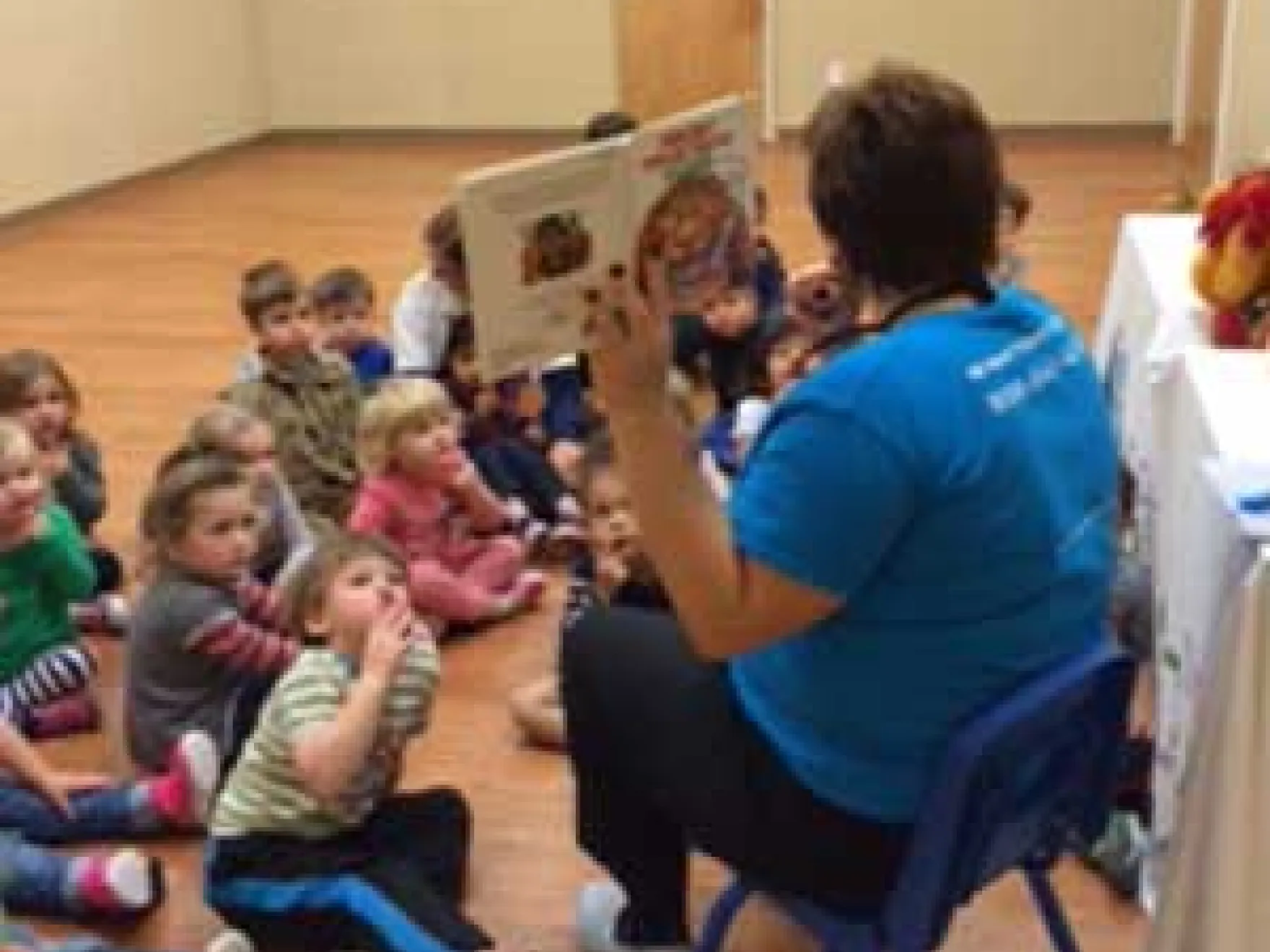 Dental professional in blue shirt reading book to group of children seated on wooden floor indoors.