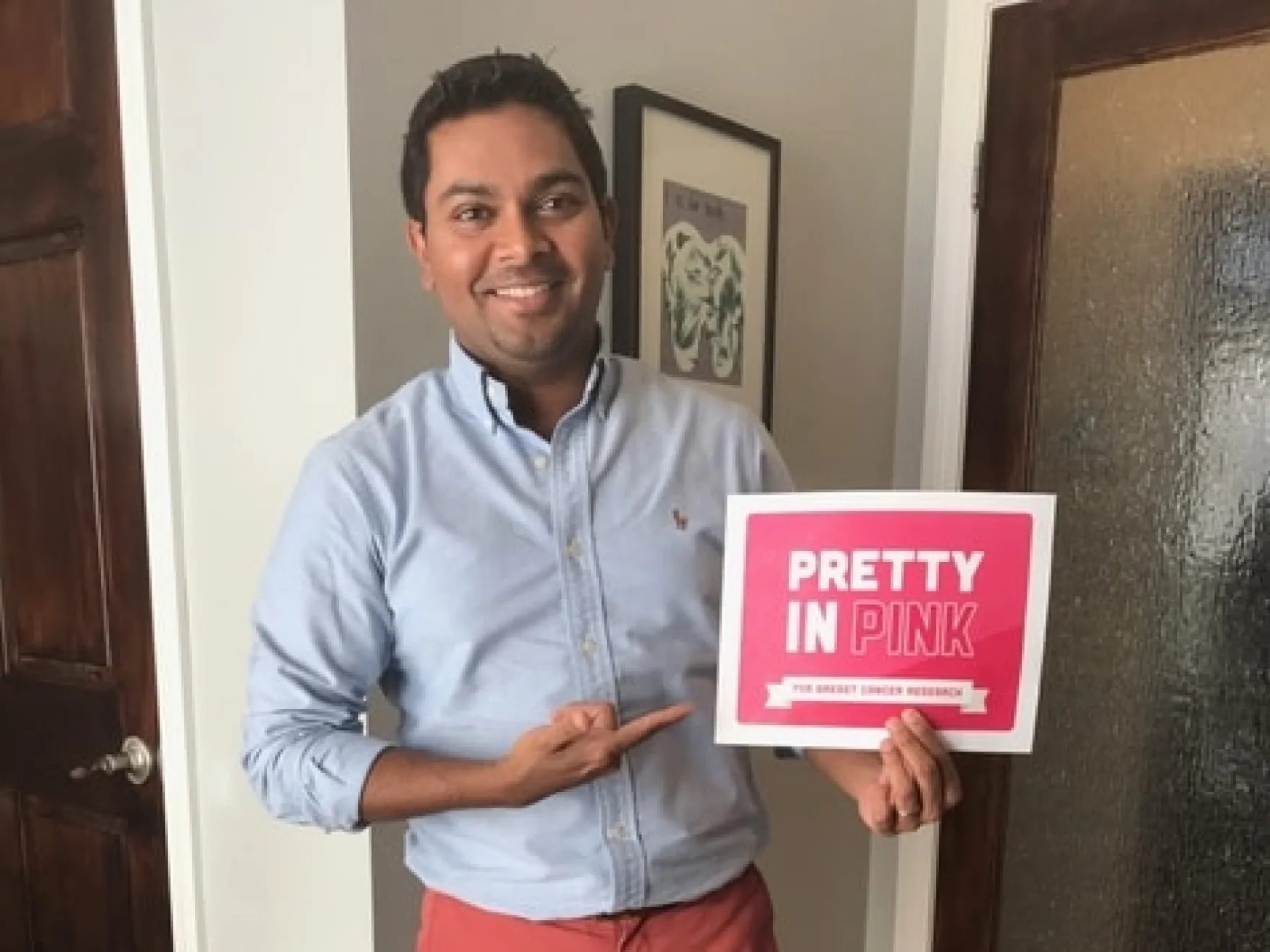 Man in blue shirt holding "Pretty in Pink" sign in hallway, smiling at camera.