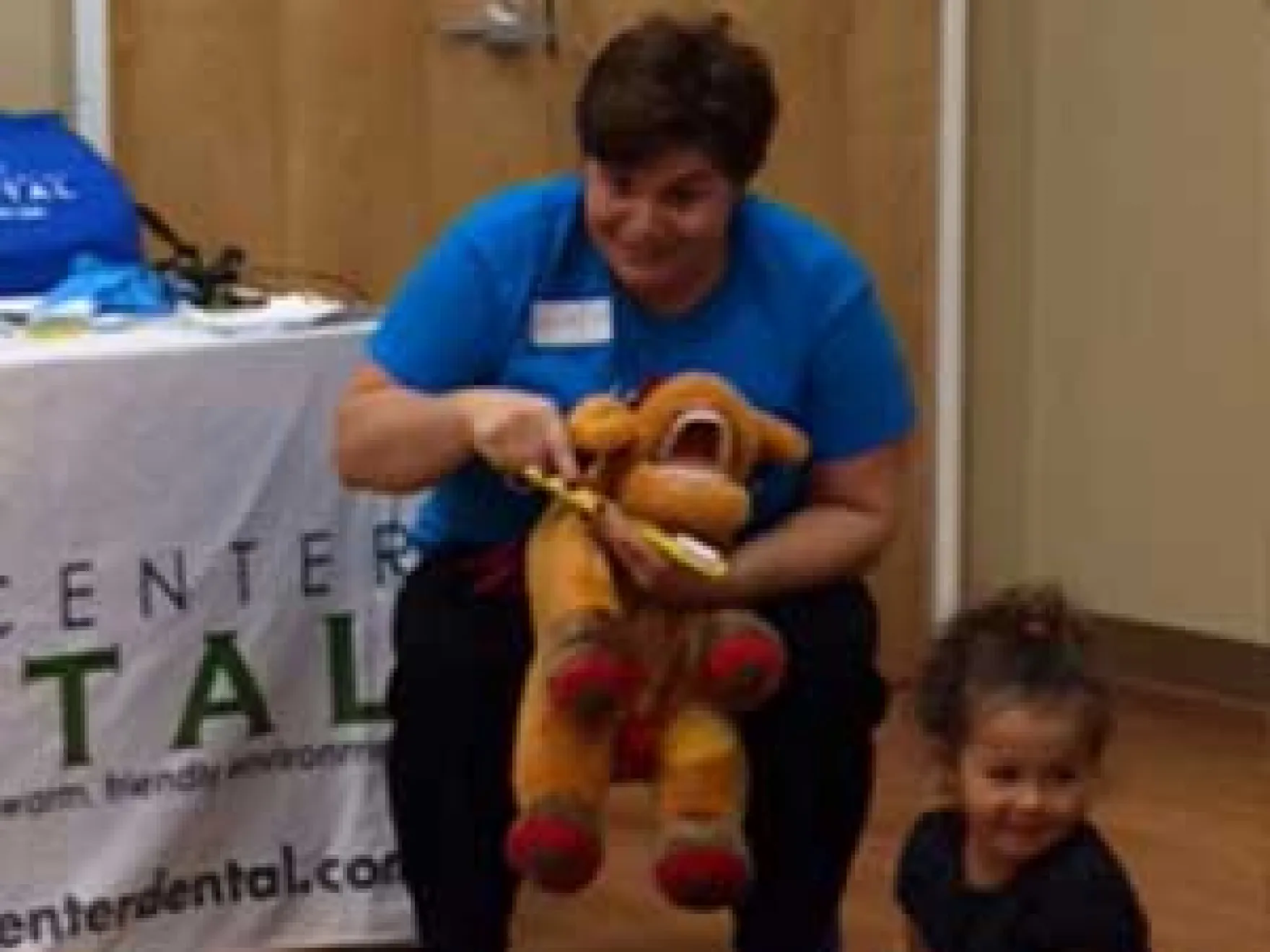 Dental professional in blue shirt demonstrating puppet toy to young child at dental office event.