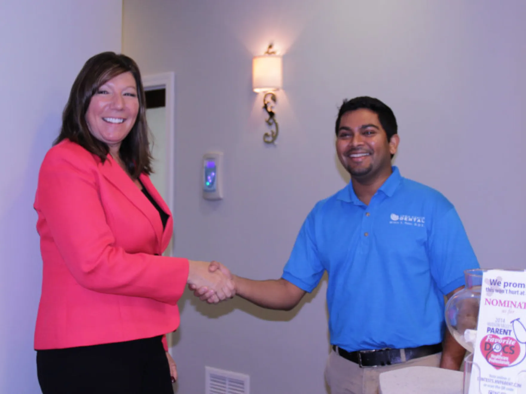 Two professionals shaking hands in dental office, woman in pink blazer, man in blue polo shirt.
