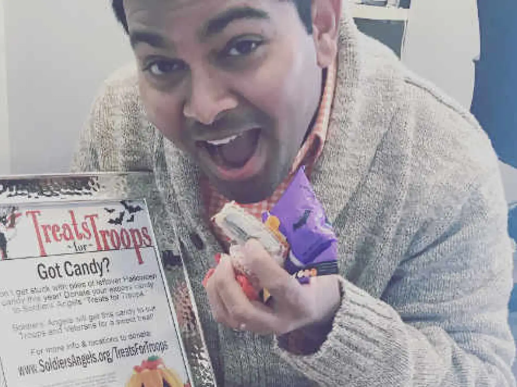 Man holding candy next to "Treats" flyer, smiling at camera indoors.