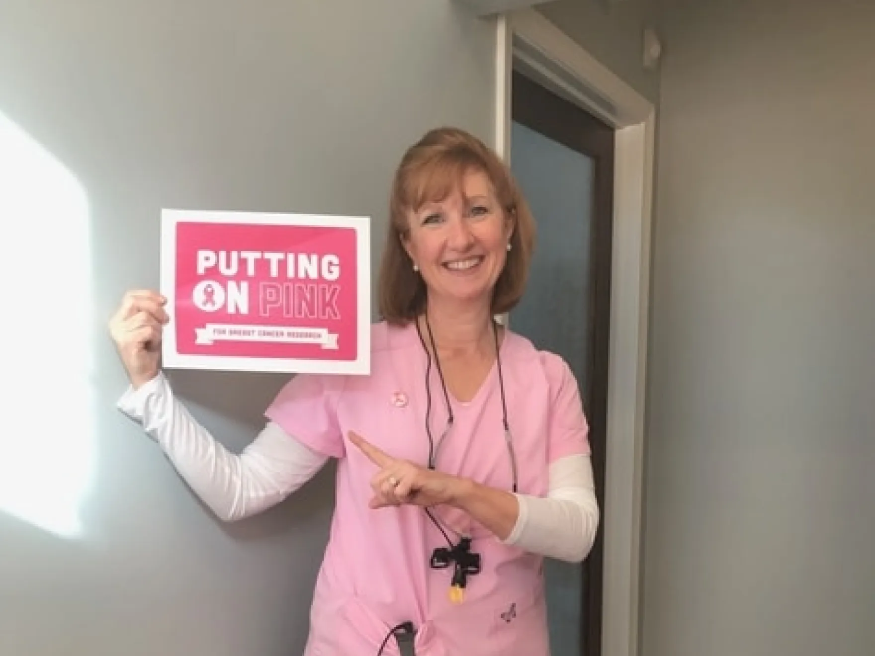 Woman in pink scrubs holding "Putting on Pink" cancer research sign indoors.