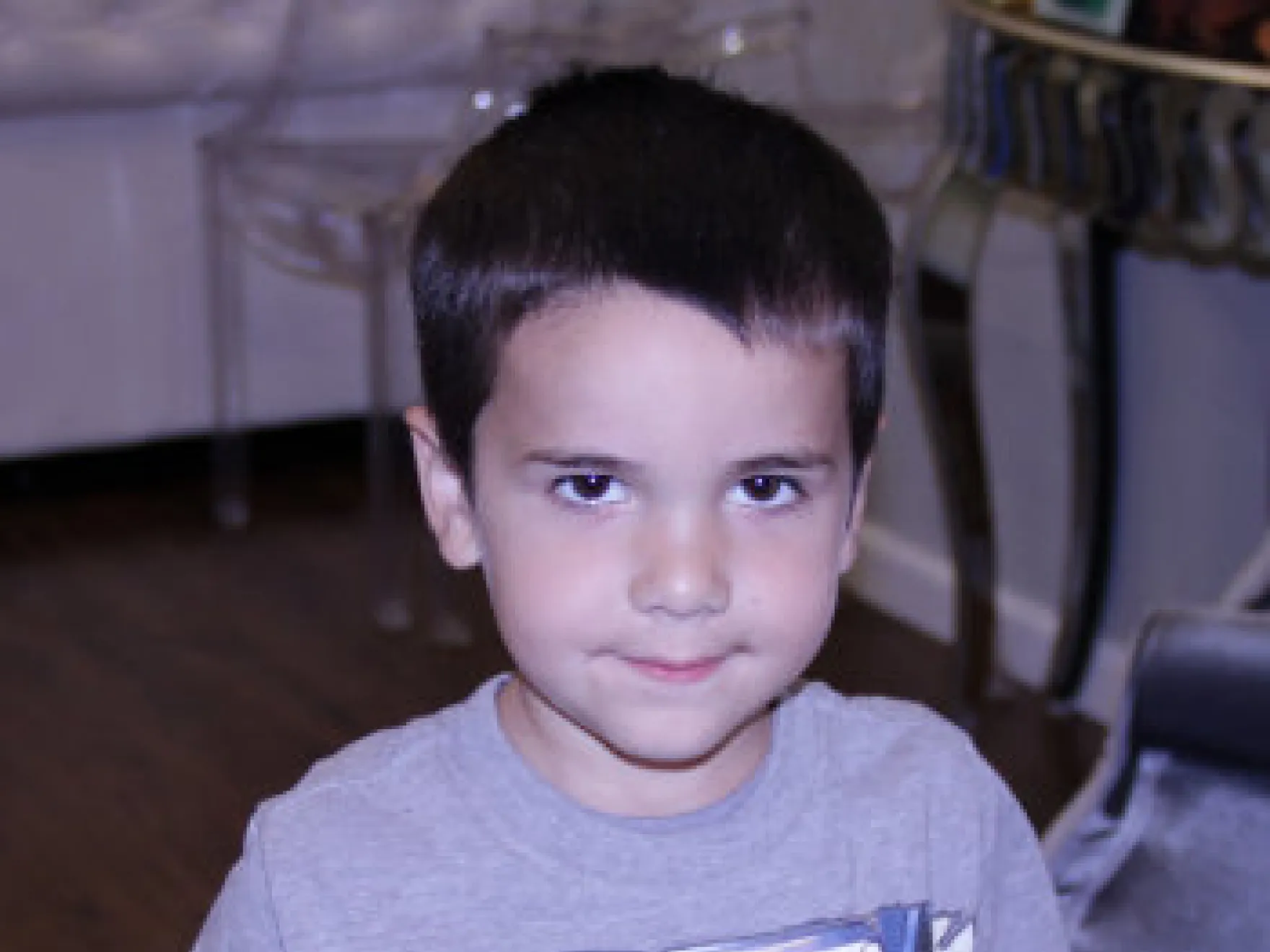 Young boy in gray shirt smiling at camera in indoor residential setting.