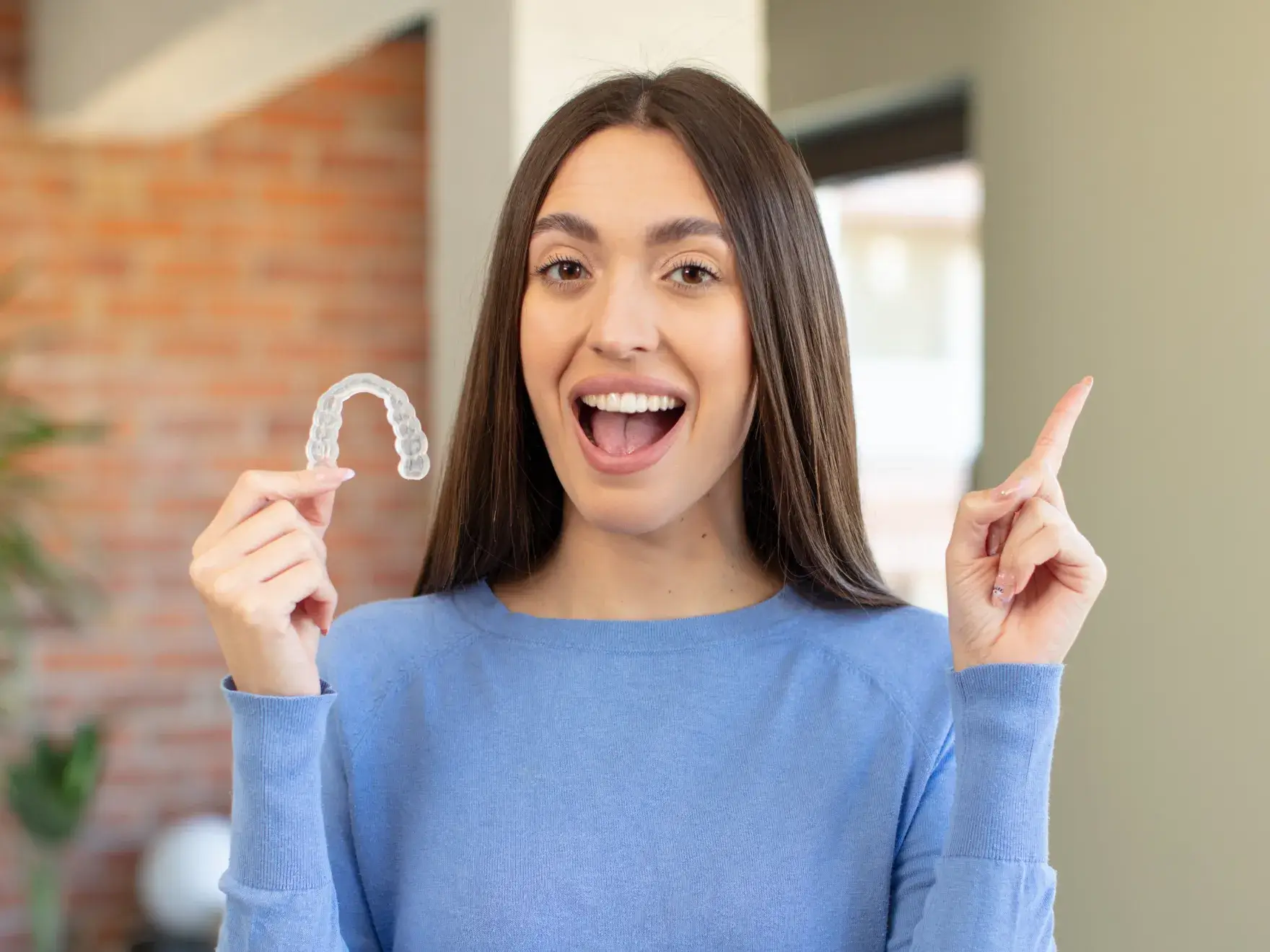 Woman holding clear aligner, smiling excitedly in modern dental office setting.