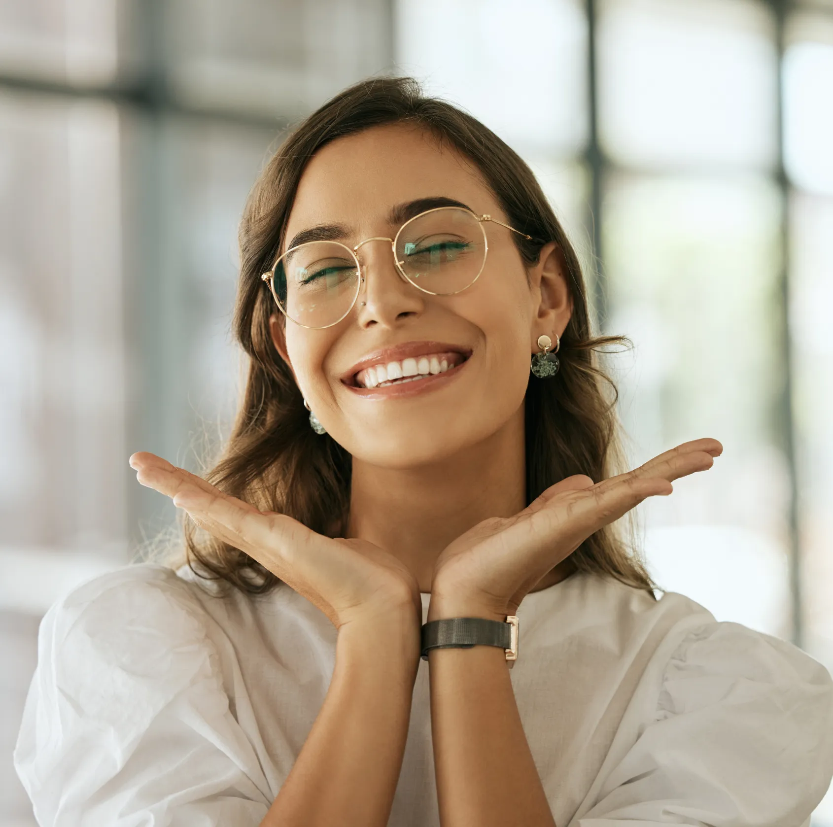 Happy woman wearing glasses smiling with hands cupped near face in bright office setting.