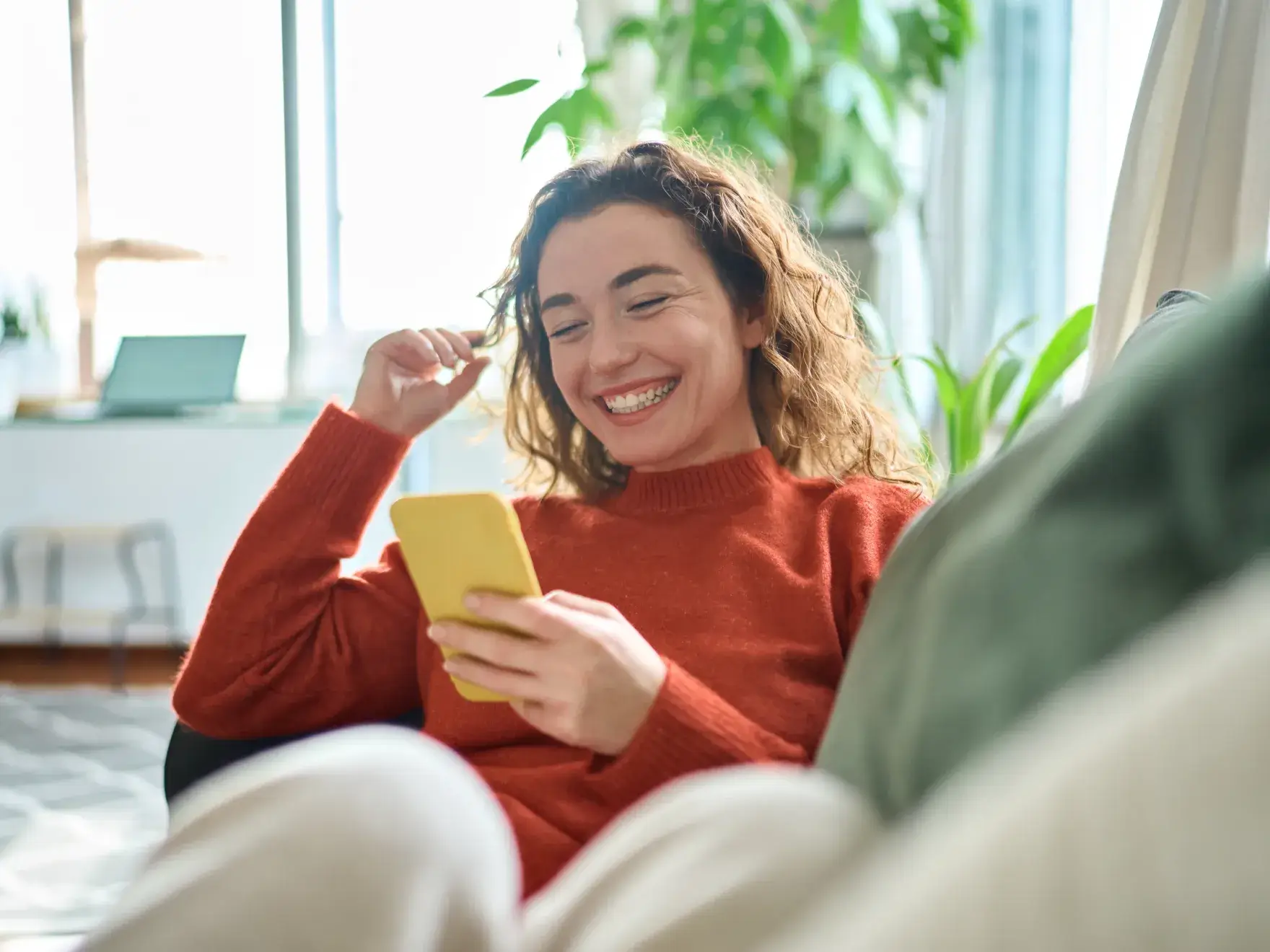 Smiling woman in red sweater holding yellow phone in bright, plant-filled room.