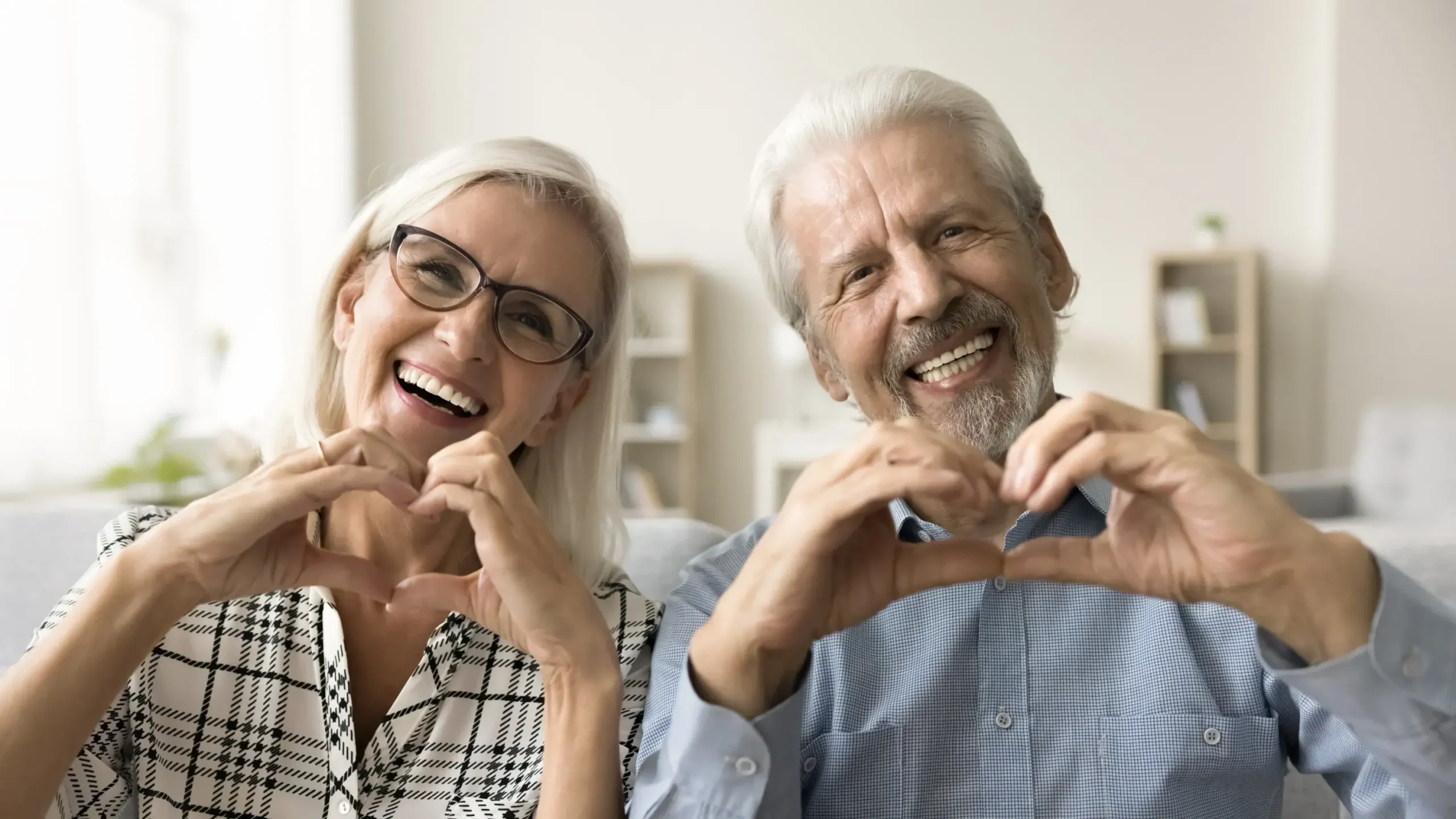 Happy mature couple making heart hand gesture in bright dental office setting.