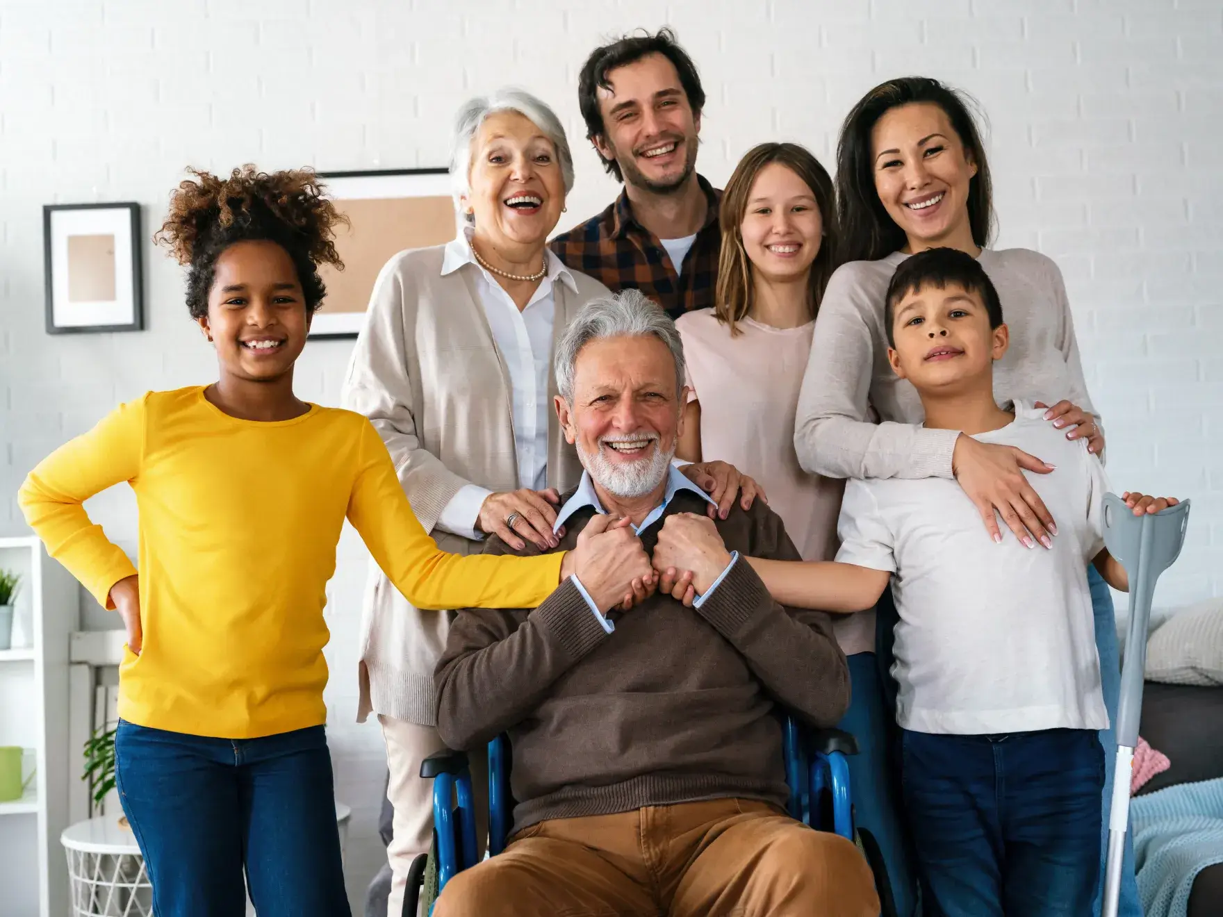 Multigenerational family smiling together indoors, including elderly man in wheelchair surrounded by loved ones.