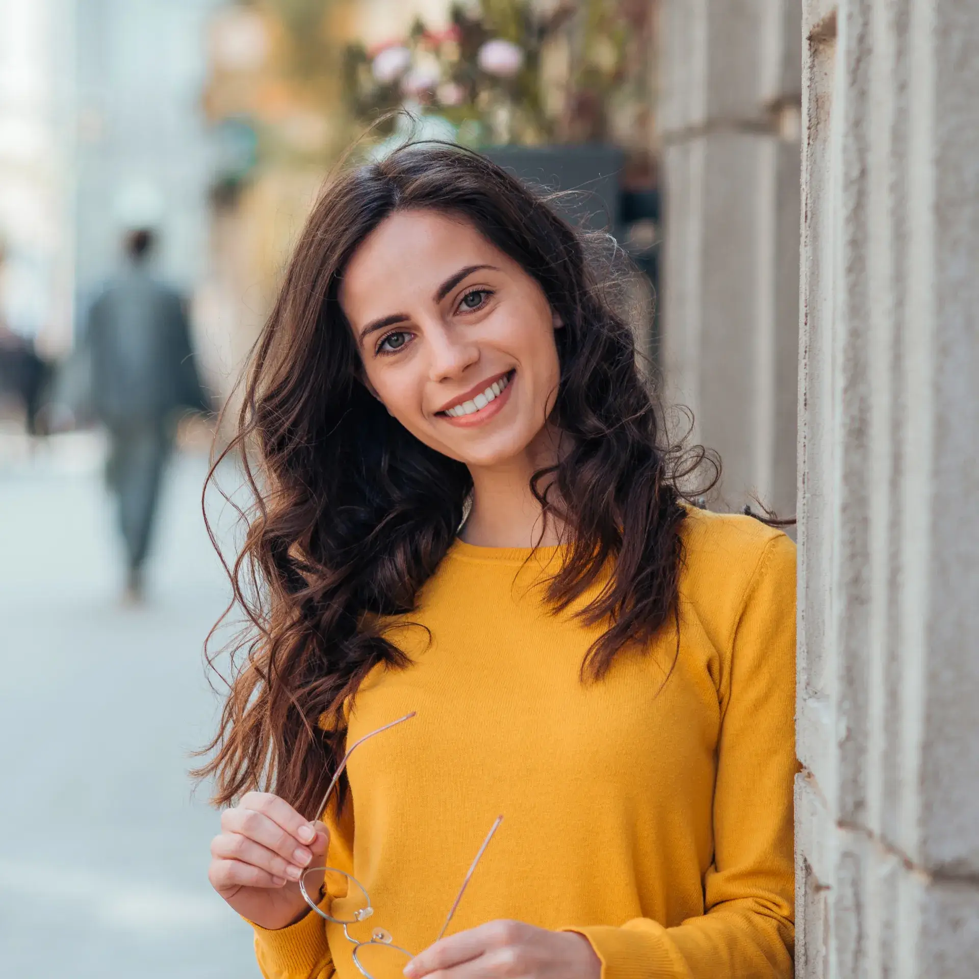 Woman in yellow sweater smiling against brick wall in urban street setting.