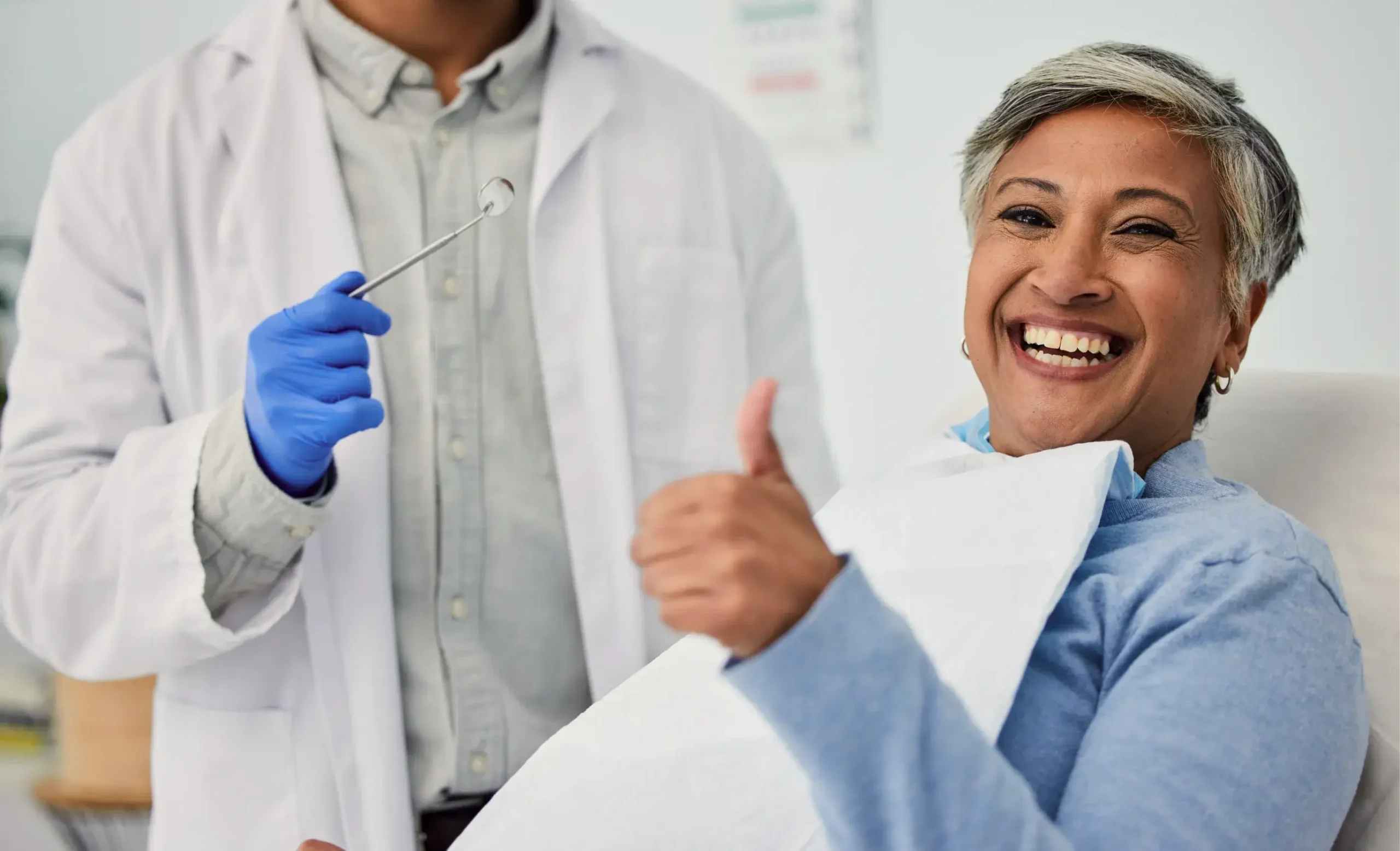 Smiling patient giving thumbs up during dental examination with dentist holding mirror in clinical setting.