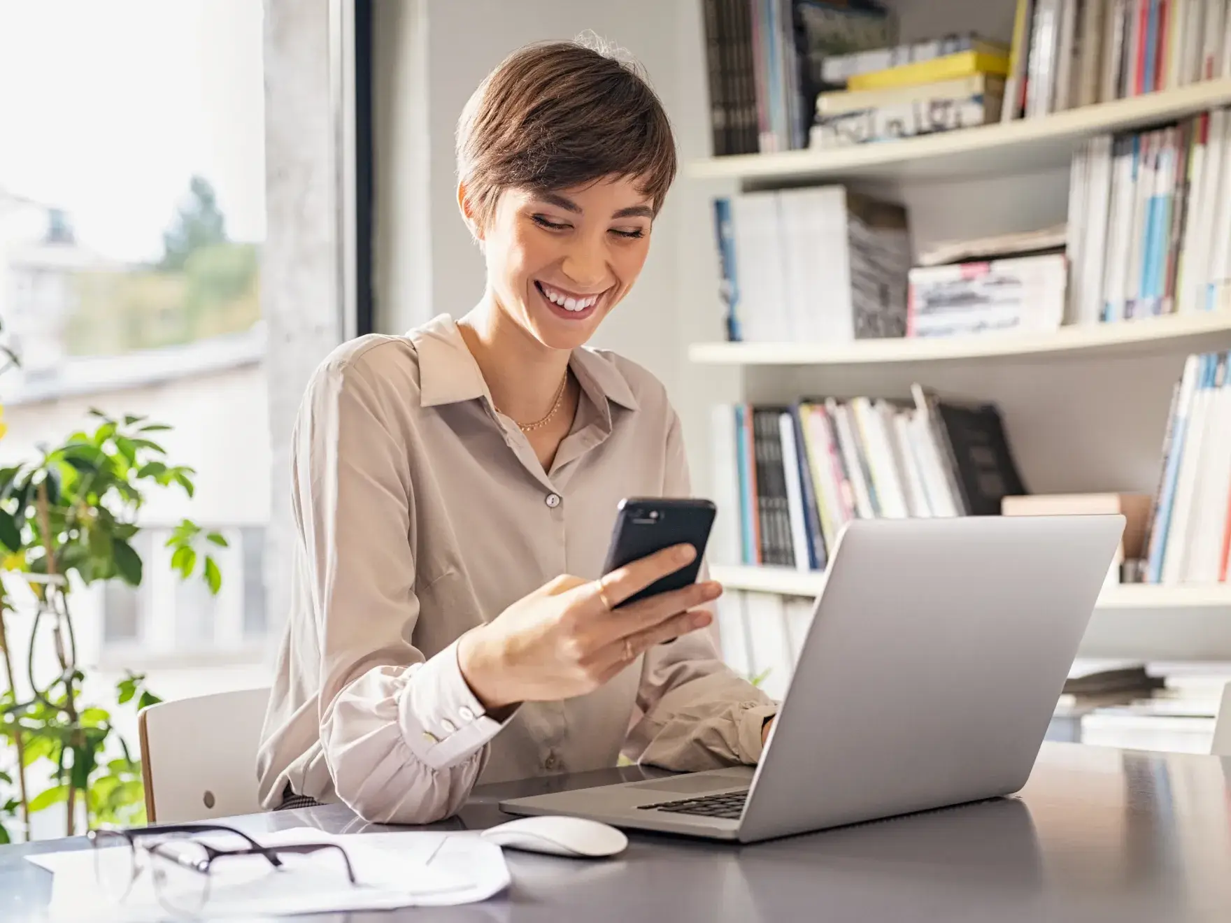 Young professional woman smiling while using smartphone at desk with laptop in modern office.