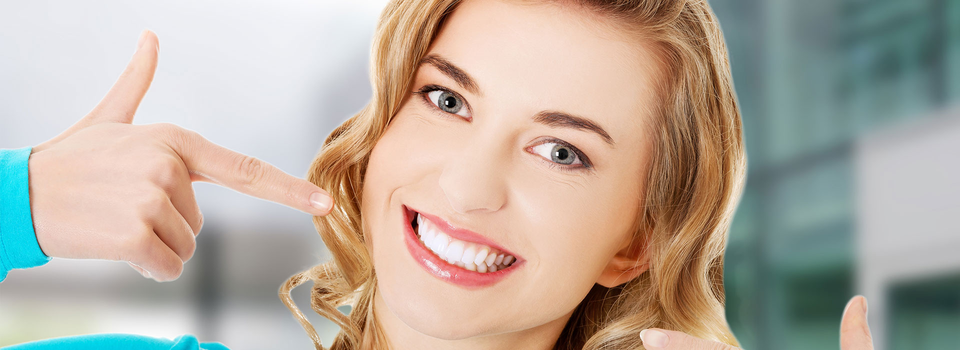 Woman pointing to her bright smile wearing turquoise top against blurred background.