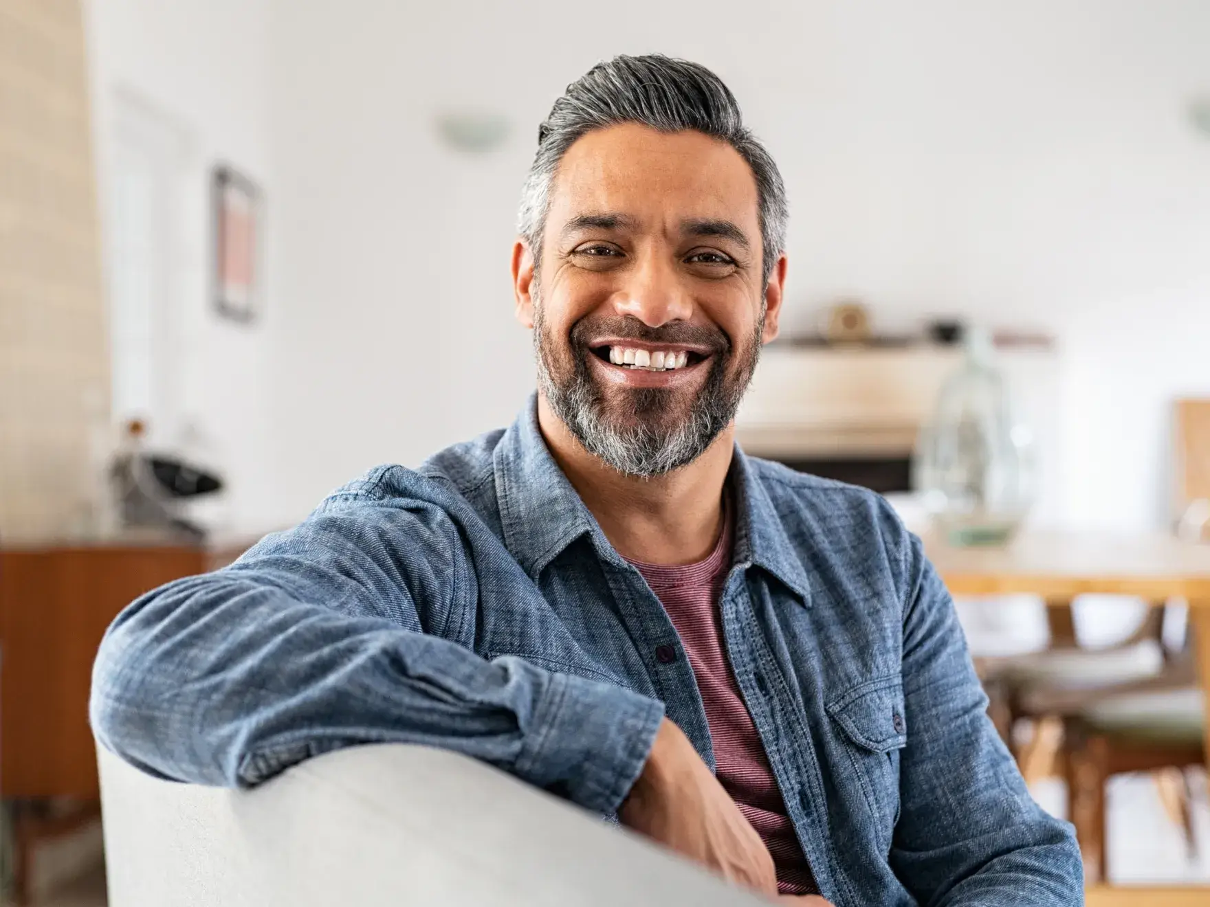 Smiling man in denim shirt relaxing on couch in modern dental office waiting room.