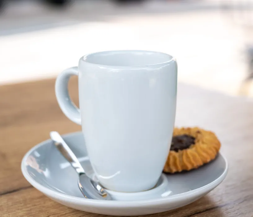 White coffee mug on saucer with spoon and cookies on wooden table.