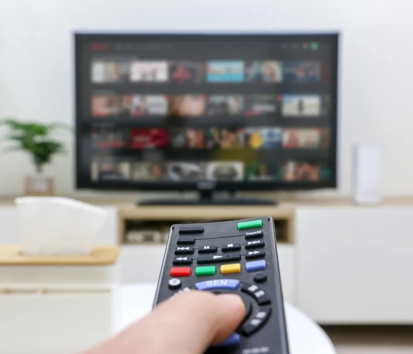 Hand holding TV remote control pointed at streaming service menu on television screen in living room.