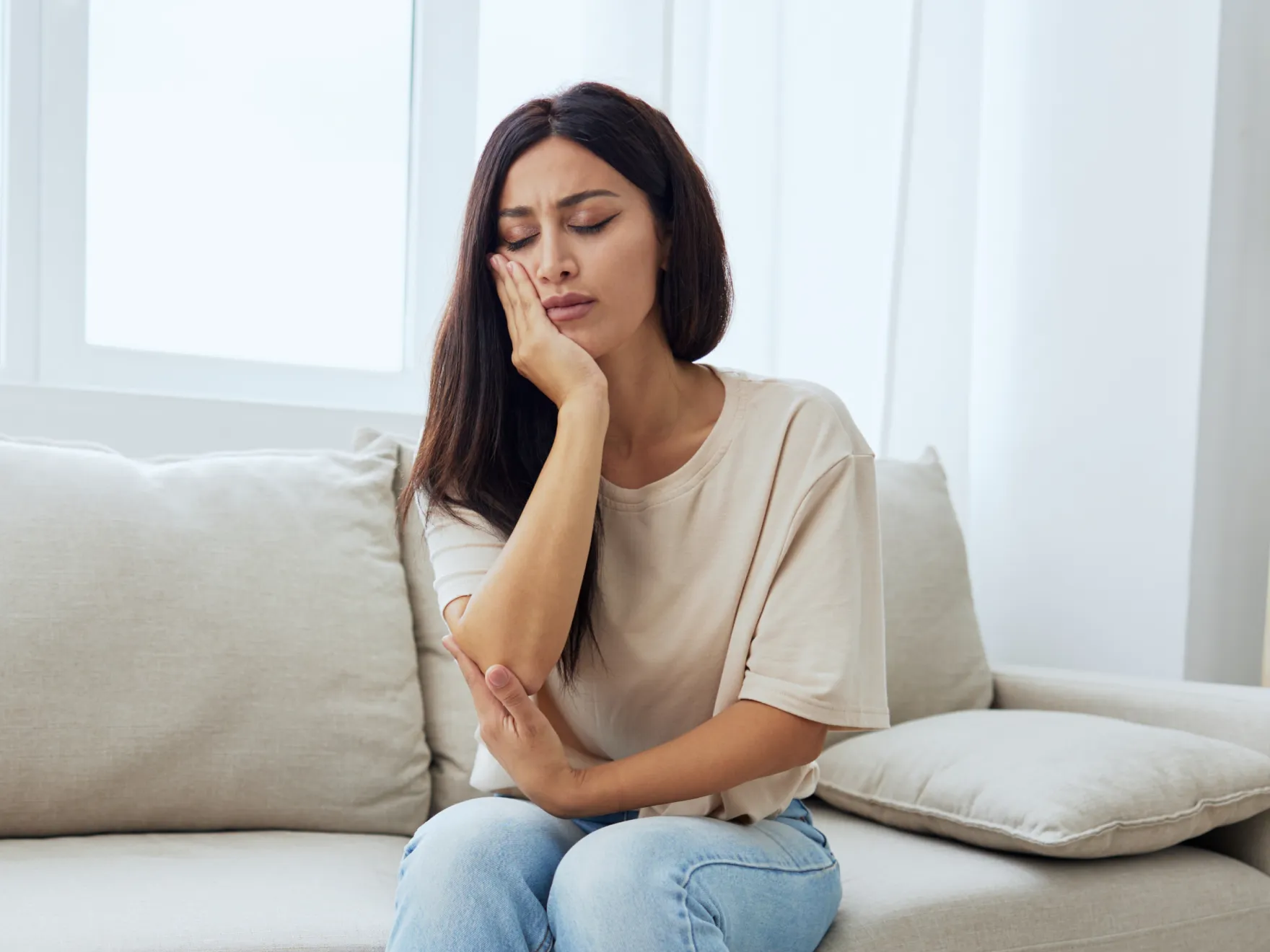 Woman with toothache holding cheek in pain, sitting on couch at home.