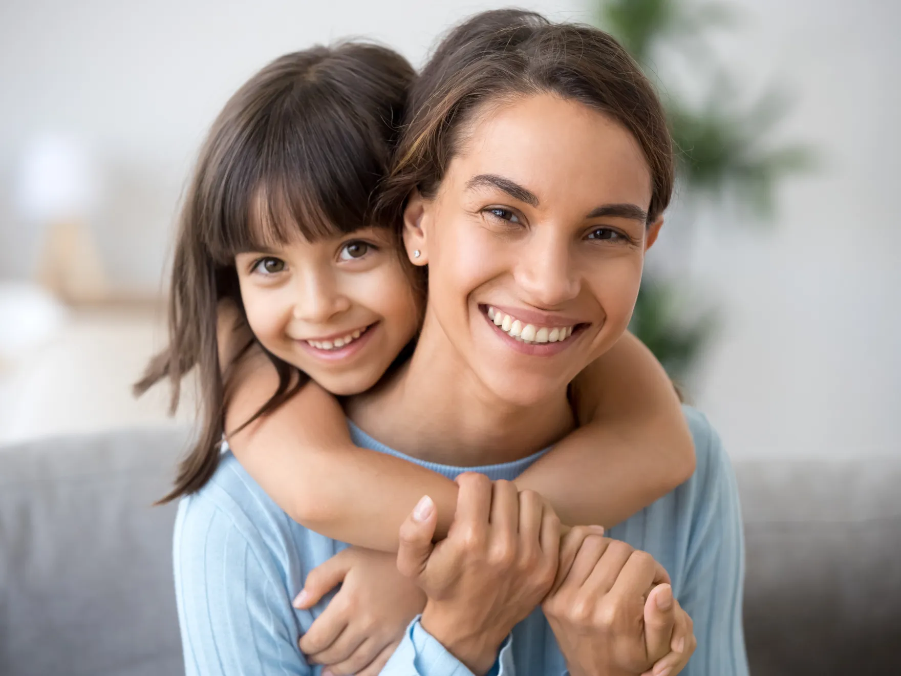 Woman holding young girl on shoulders, both smiling at camera in bright home setting.