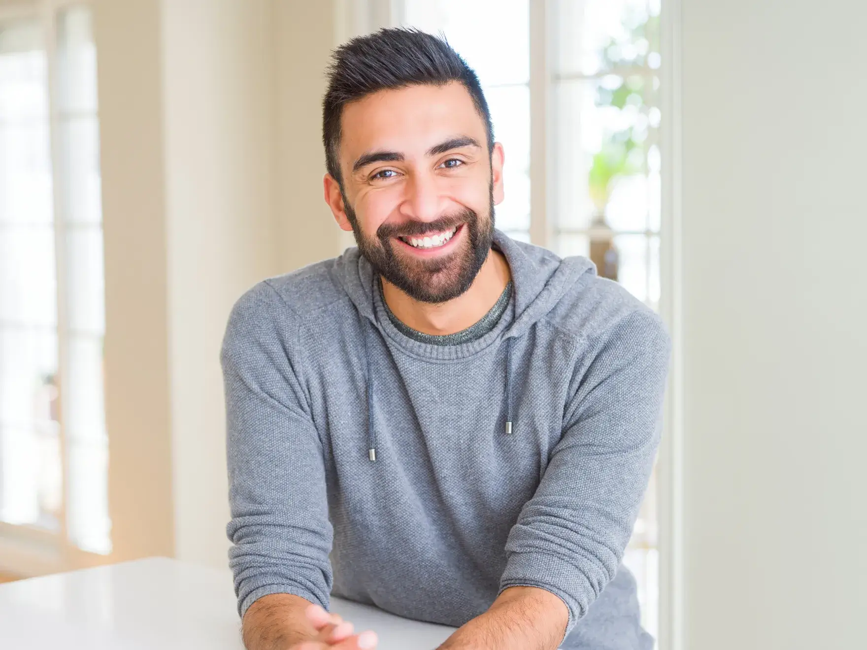 Smiling man in gray sweater leaning on table in bright, modern dental office setting.