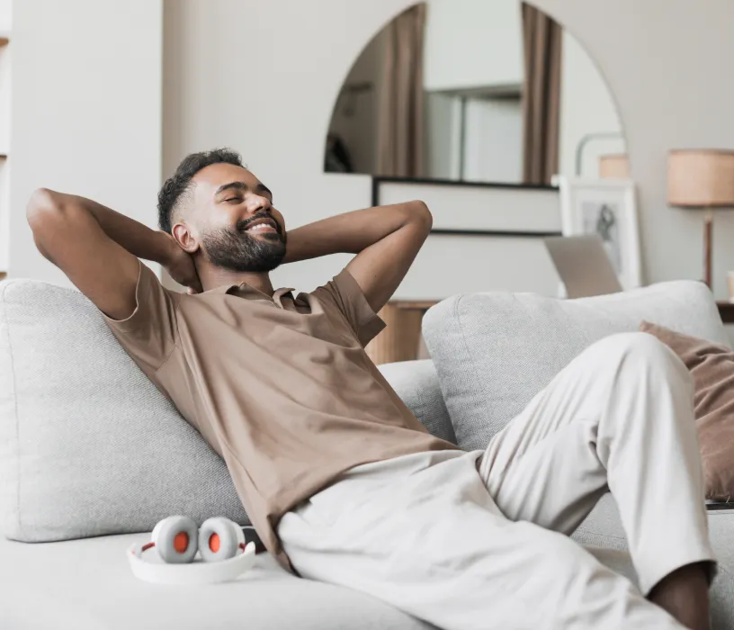 Man relaxing on couch at home with hands behind head, smiling contentedly.