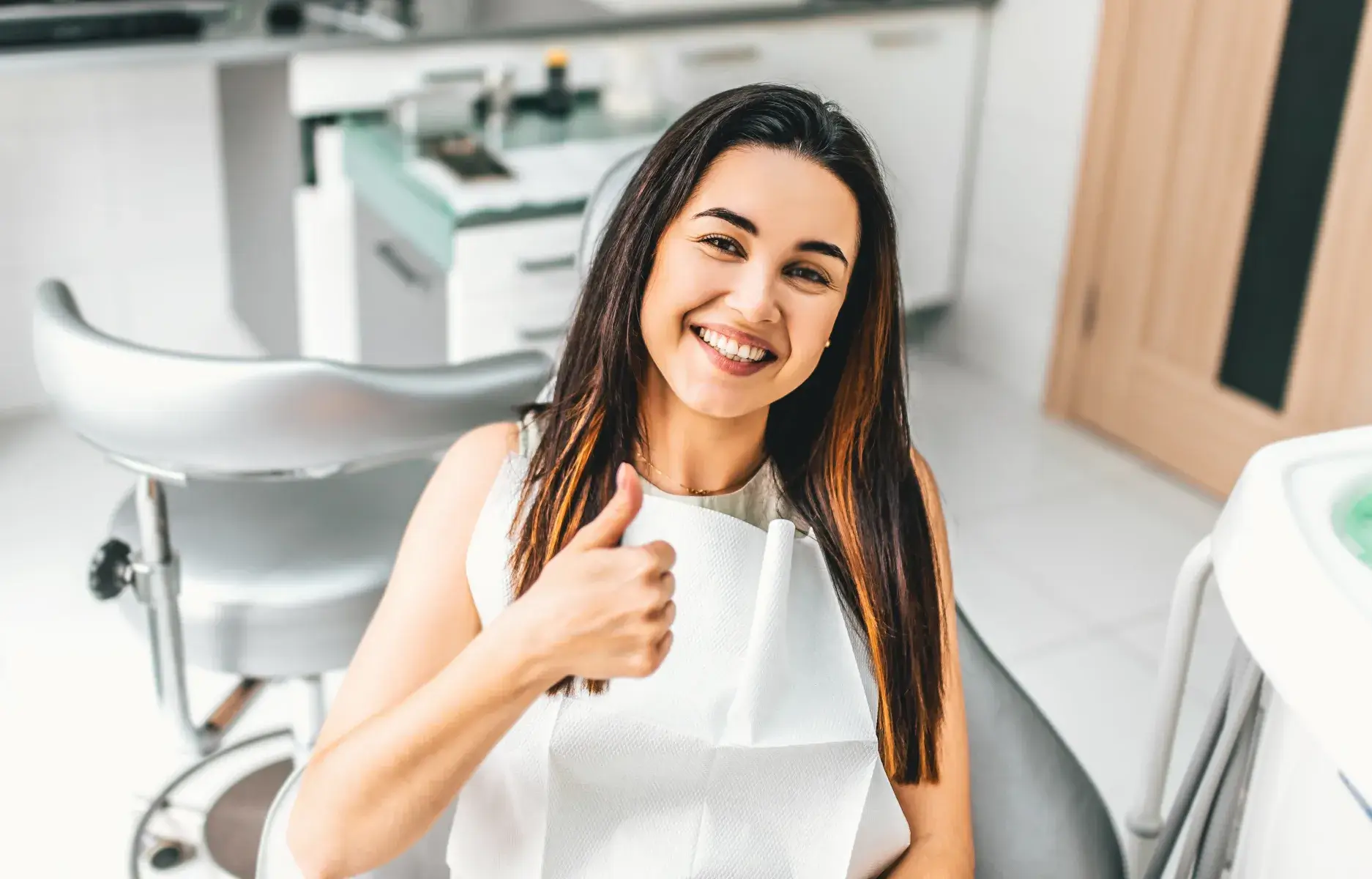 Smiling woman giving thumbs up in modern dental office treatment room.