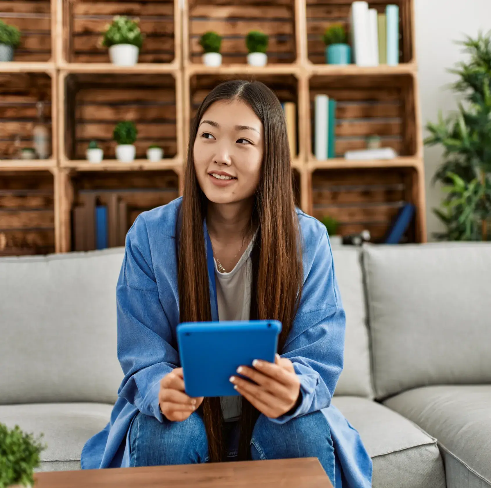 Young woman smiling while holding blue tablet, sitting on gray couch in modern office with wooden shelving.