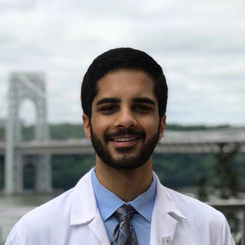 Professional male dentist in white coat and blue shirt smiling outdoors with bridge in background.