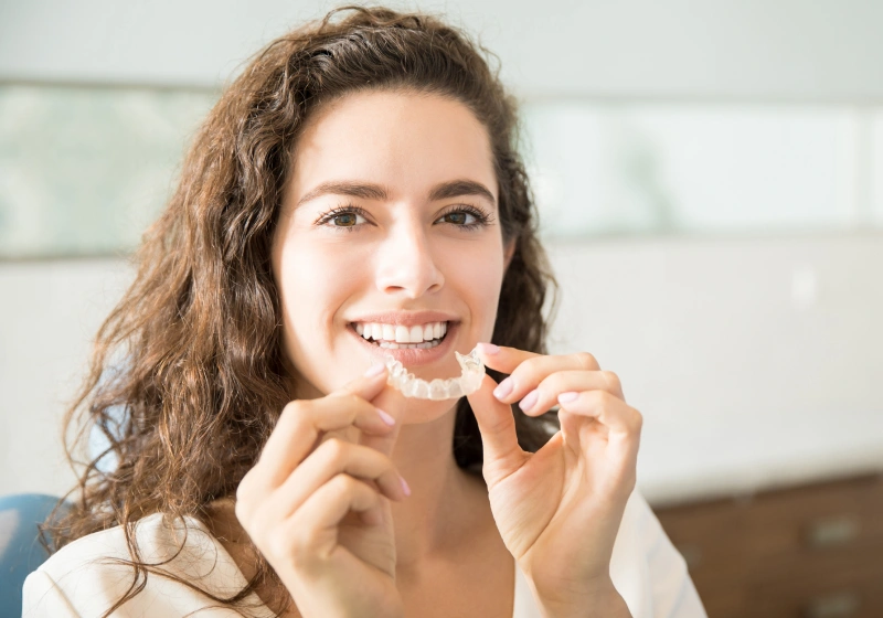 Smiling woman holding clear dental aligner in modern dental office setting.