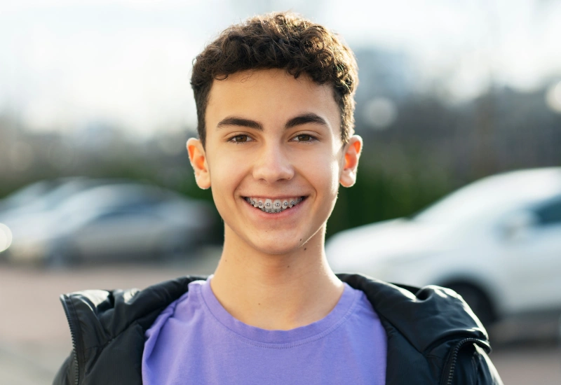 Smiling teenage boy wearing braces and purple shirt in outdoor parking lot setting.