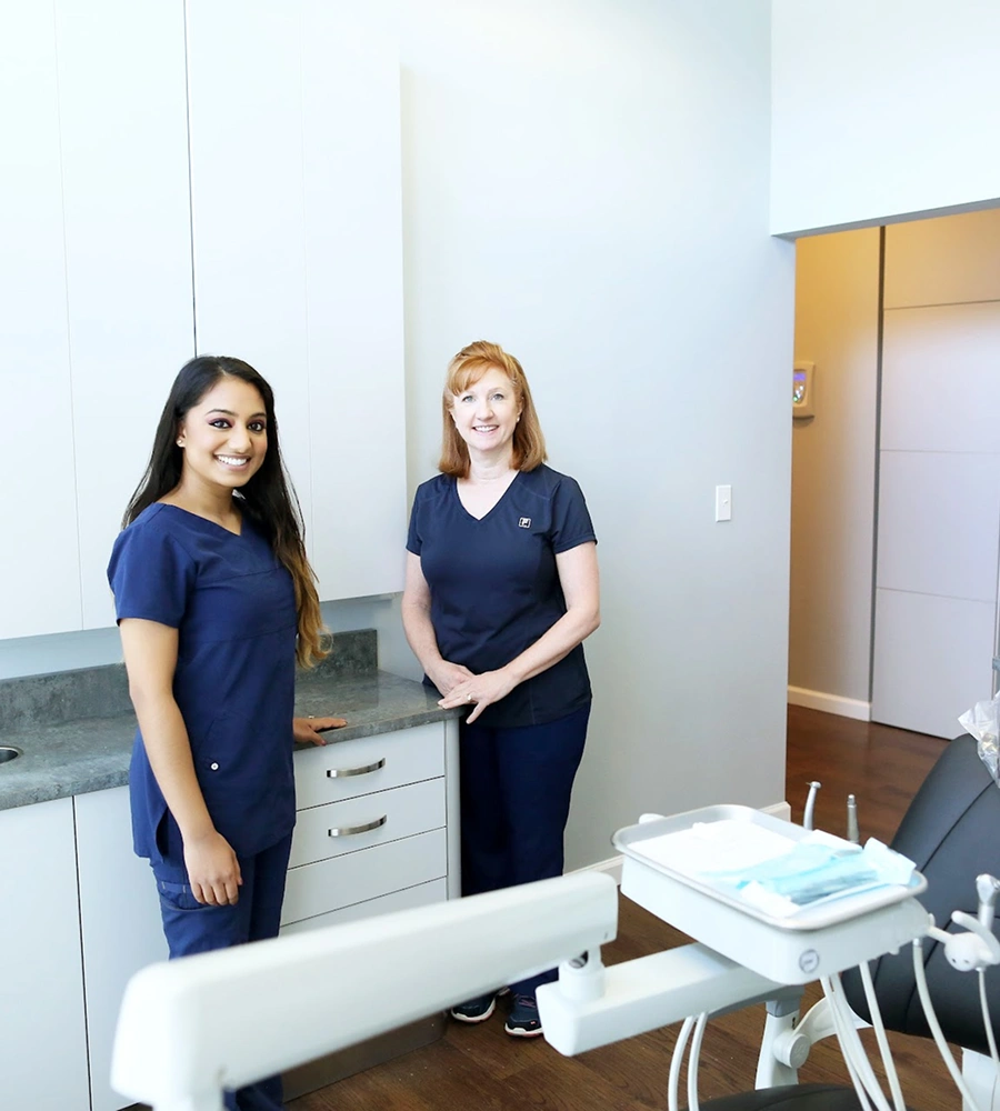 Two dental professionals in scrubs smiling in a modern dental treatment room with equipment.