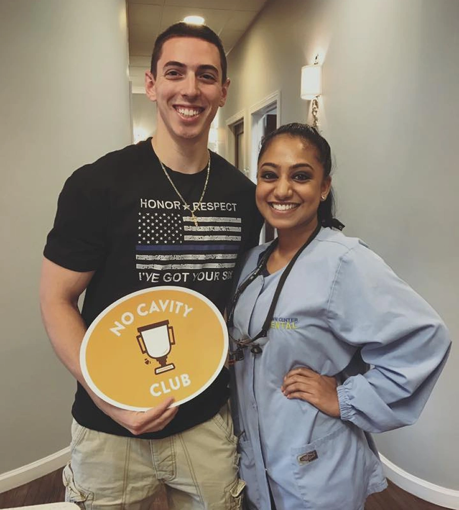 Two people smiling in dental office hallway, holding yellow "Mold Cavity Club" sign.