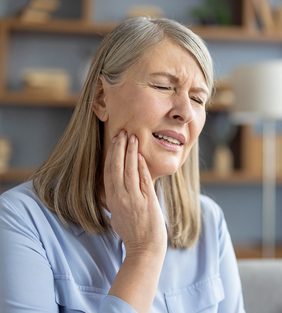 Woman experiencing tooth pain, holding jaw, in home setting wearing light blue shirt