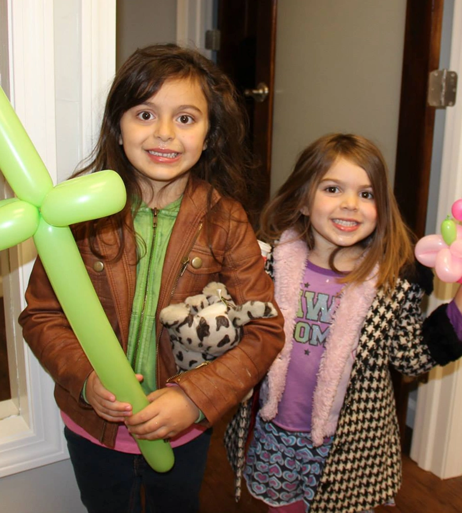 Two young girls smiling at camera holding green balloon animals at indoor celebration event.